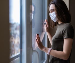 Sad young woman in a medical mask looks out of the window. Quarantine during the coronavirus pandemic.